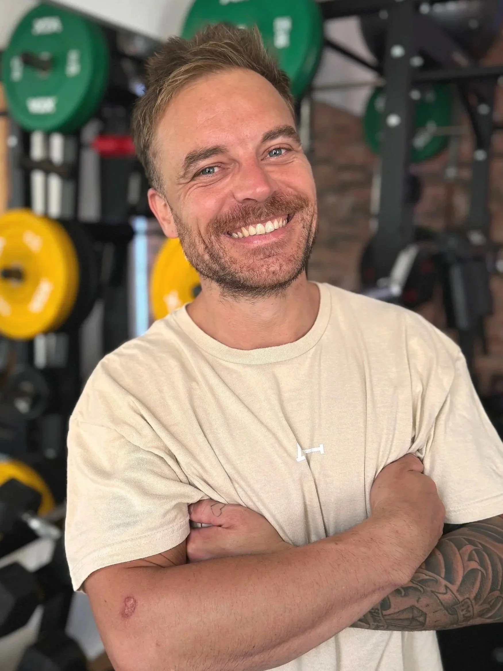 Ein lachender Mann mit Tattoos auf dem linken Arm, trägt ein beige T-Shirt, steht in einem Fitnessstudio vor Gewichten.