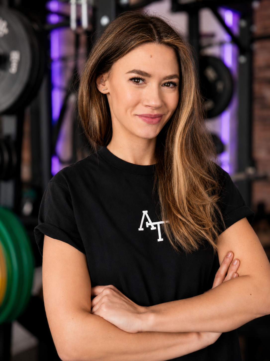 Junge Frau mit braunen Haaren in einem Fitnessstudio, lächelnd, trägt ein rotes T-Shirt mit weißen Buchstaben, vor einer Hantelbank gestanden.