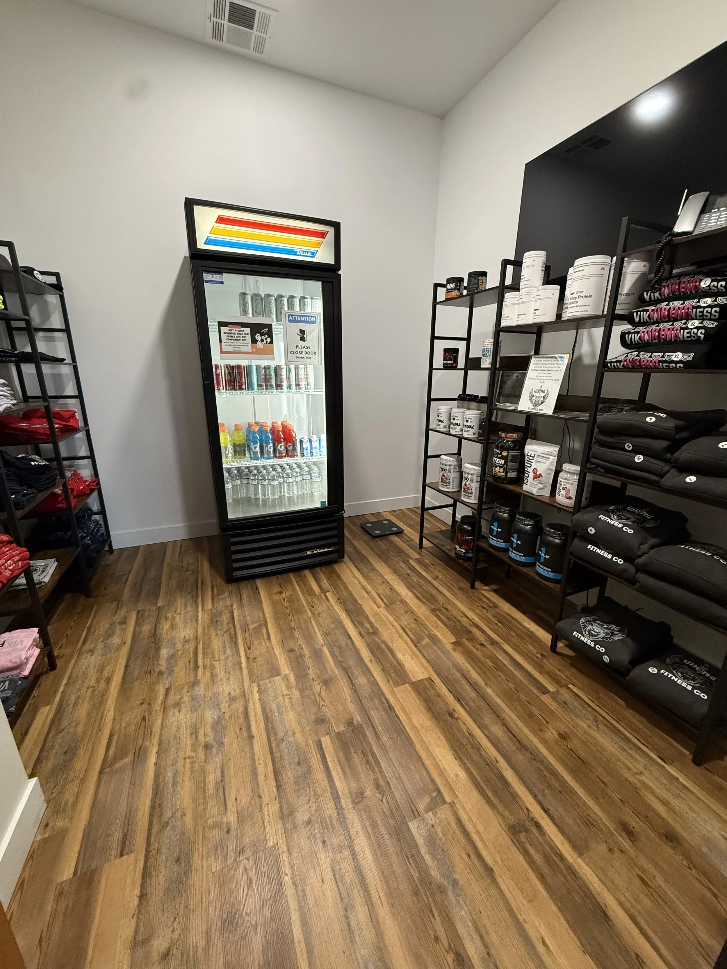 View of a retail store with wooden flooring, black shelving units displaying fitness apparel and supplements, and a glass door refrigerator stocked with beverages.