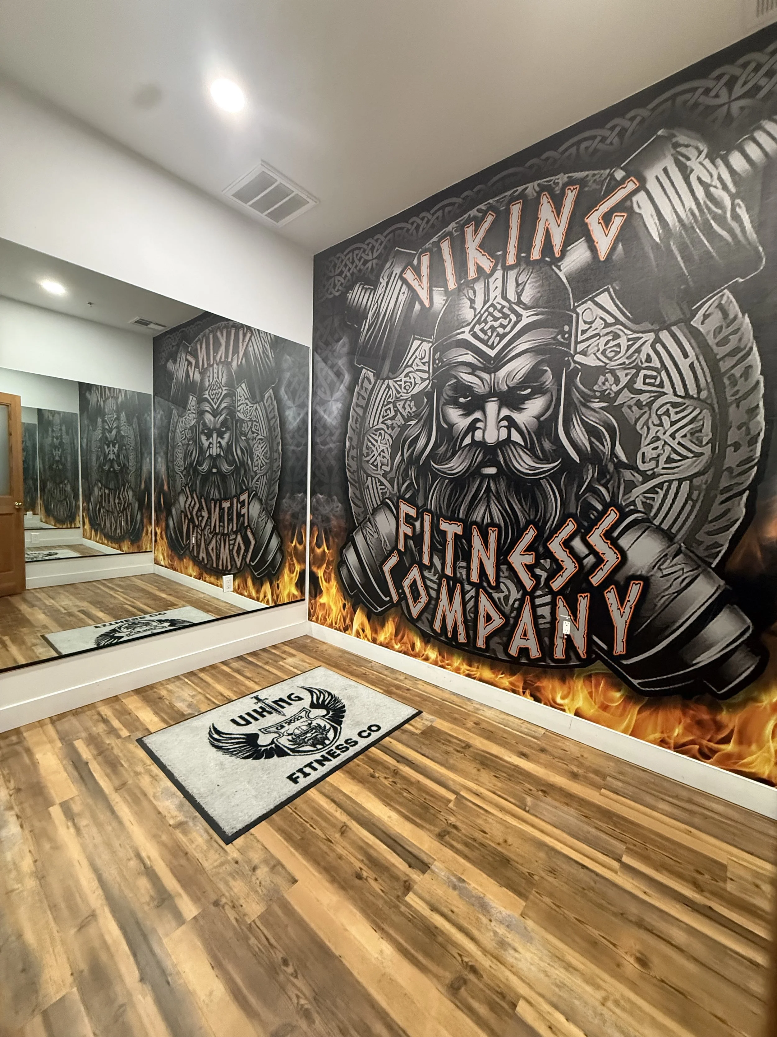 Mirror with a large Viking-themed graphic and text promoting a fitness company, flames at the bottom, and two rugs on a wooden floor.