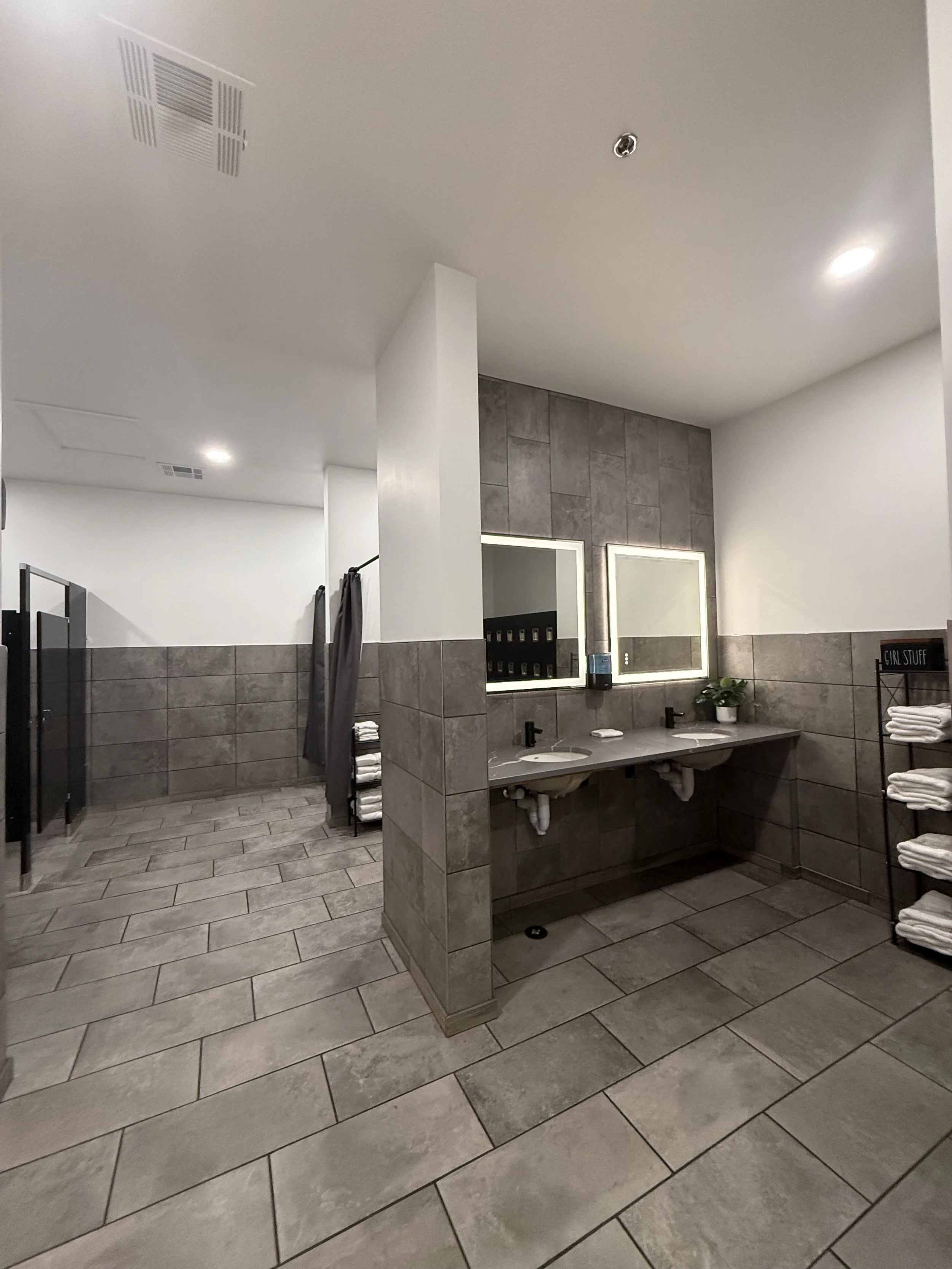 Modern bathroom with double sinks, large mirror with backlighting, gray tiled walls and floor, towels on rack, and potted plant near the sinks.