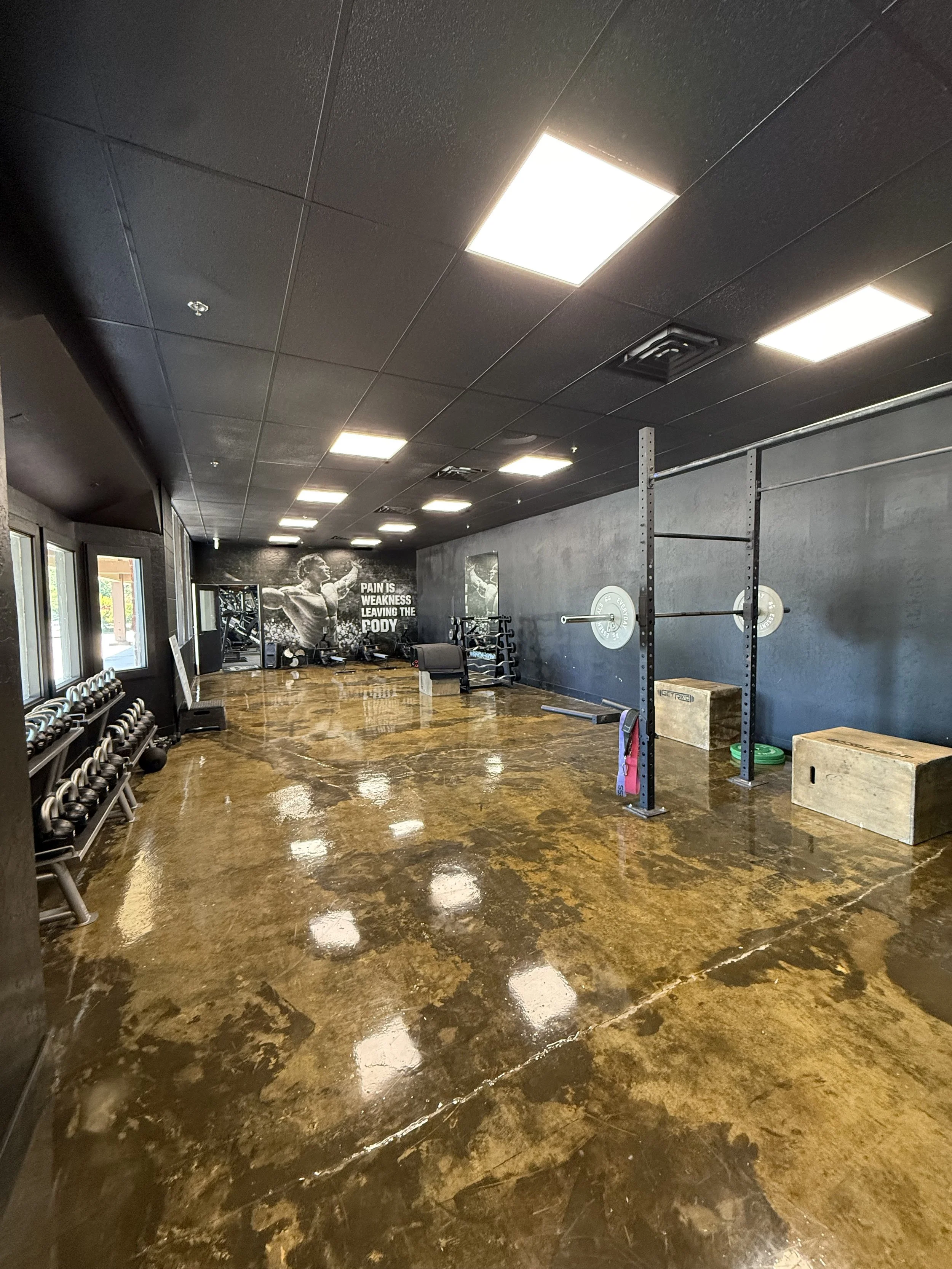 Interior of a gym with black walls, mirrors, and gym equipment including kettlebells, weight racks, and plyometric boxes. Polished brown marble flooring reflects the overhead fluorescent lights.