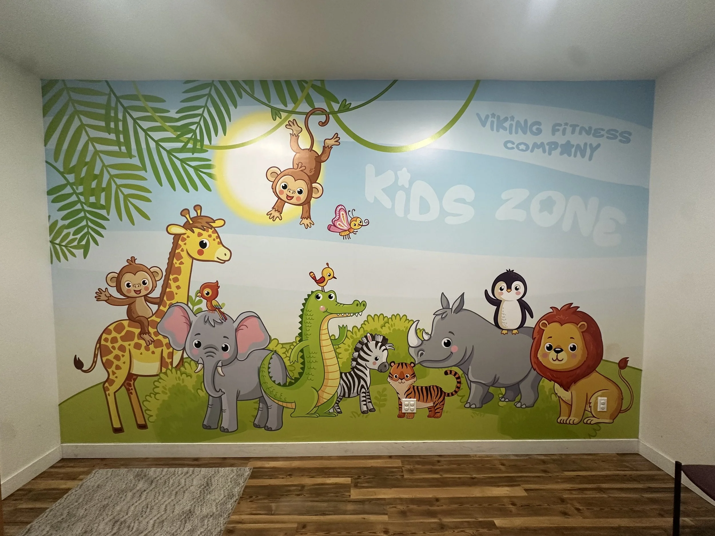 Colorful cartoon mural of jungle animals including a giraffe, elephant, crocodile, zebra, lion, monkey, penguin, tiger, and others, with green foliage and blue sky background, in a kids' zone at Viking Fitness.