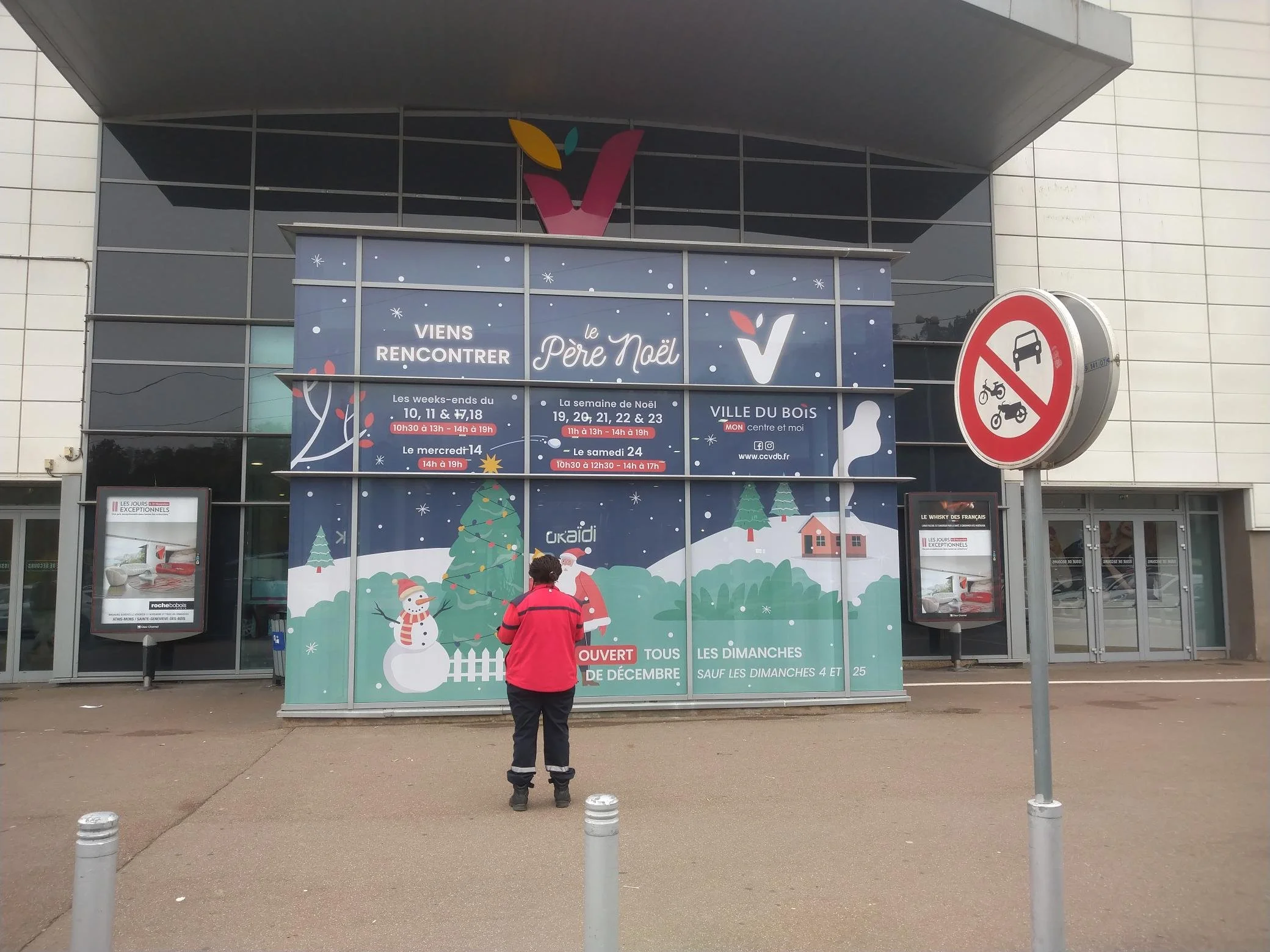 Façade d'un centre commercial avec affichage publicitaire pour Noël, un homme en veste rouge regarde l'affiche, panneau de signalisation interdisant cyclomoteurs et voitures.