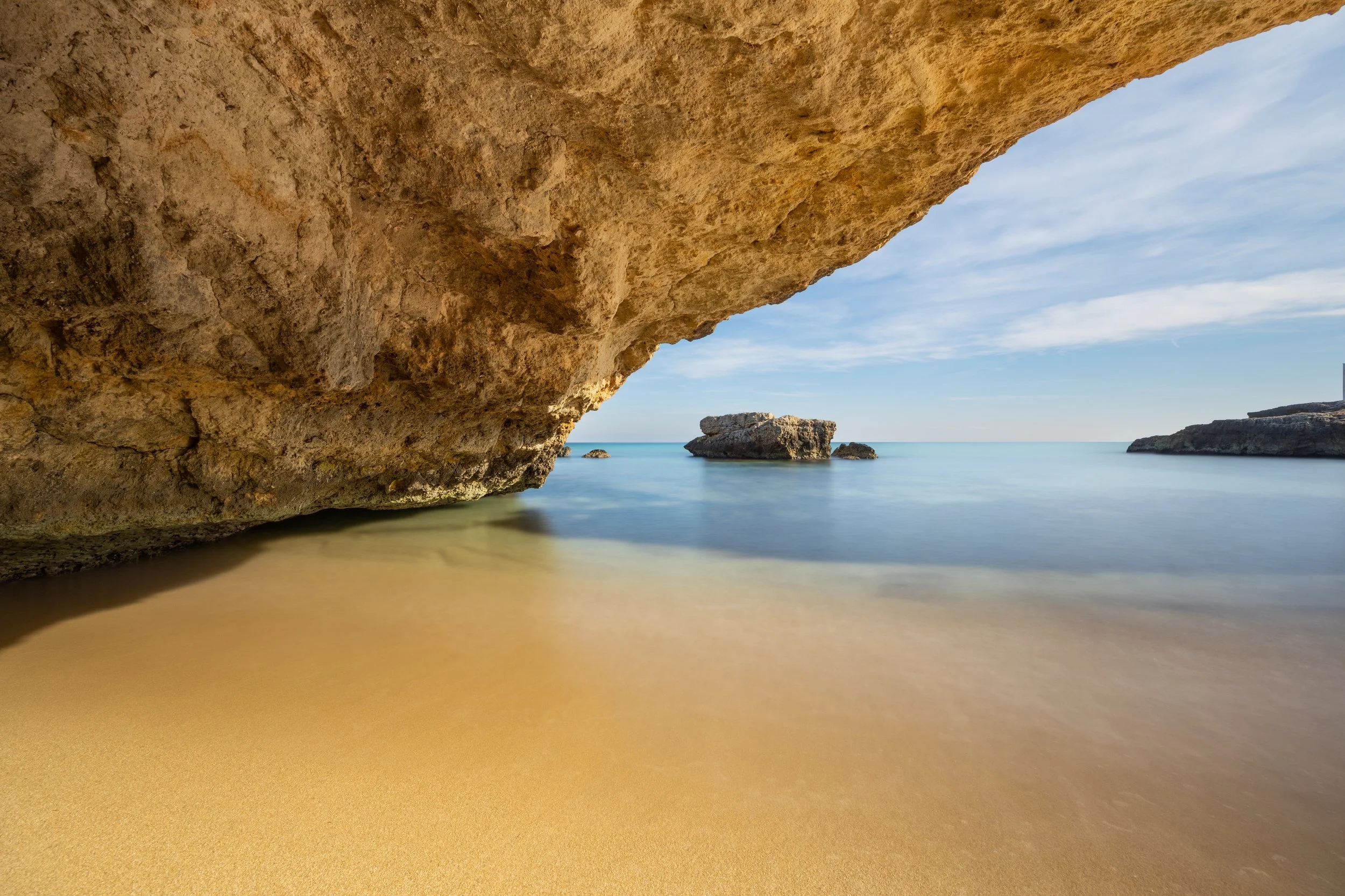 Strand mit Felsen unter einem sandfarbenen Felsen, ruhiges Wasser und blauer Himmel. Fotografie.