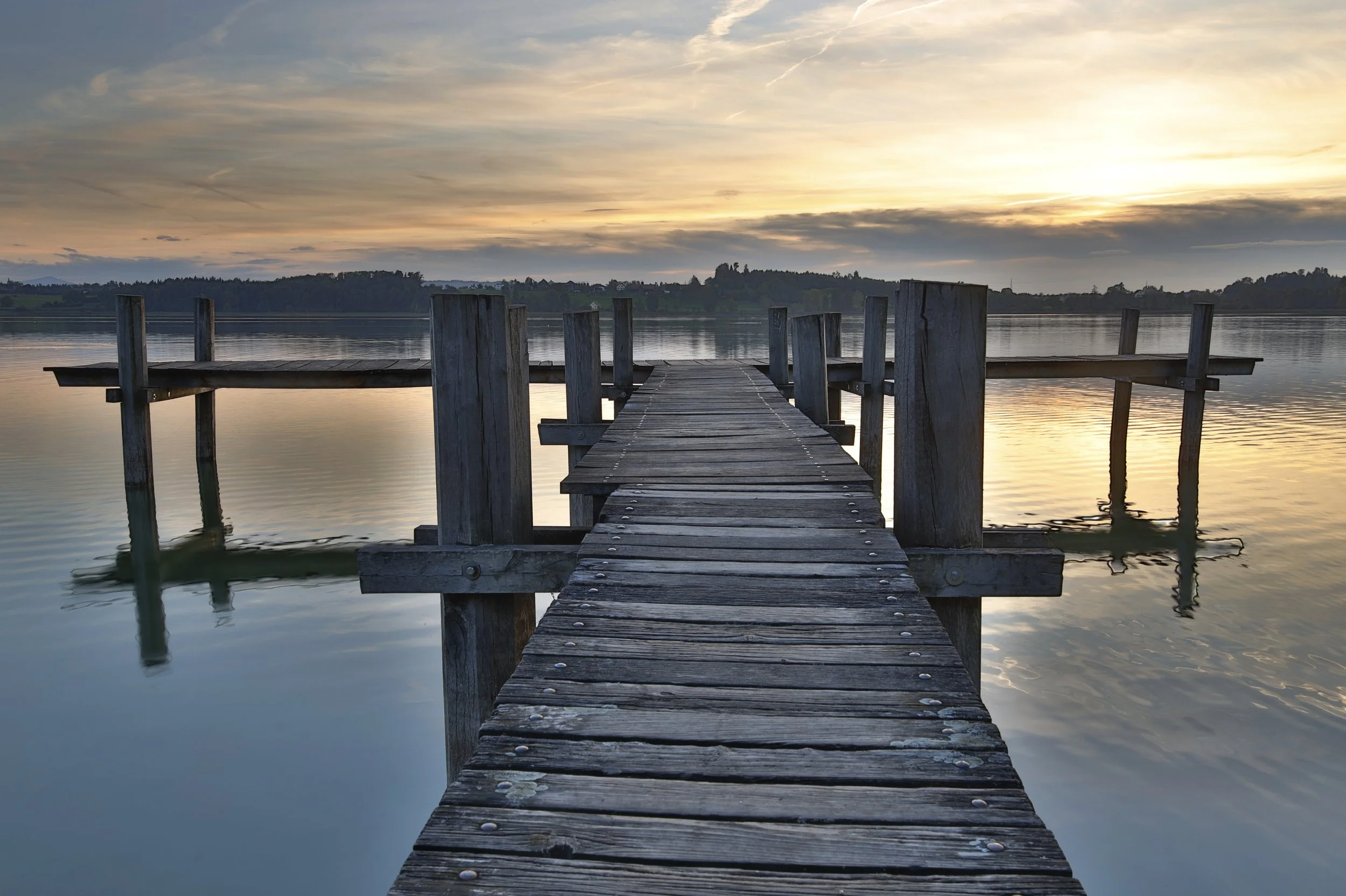 Ein Holzsteg führt ins Wasser, bei Sonnenuntergang, mit Himmel und Wasser, die ruhige Wasser reflektieren den Himmel. Fotografie.