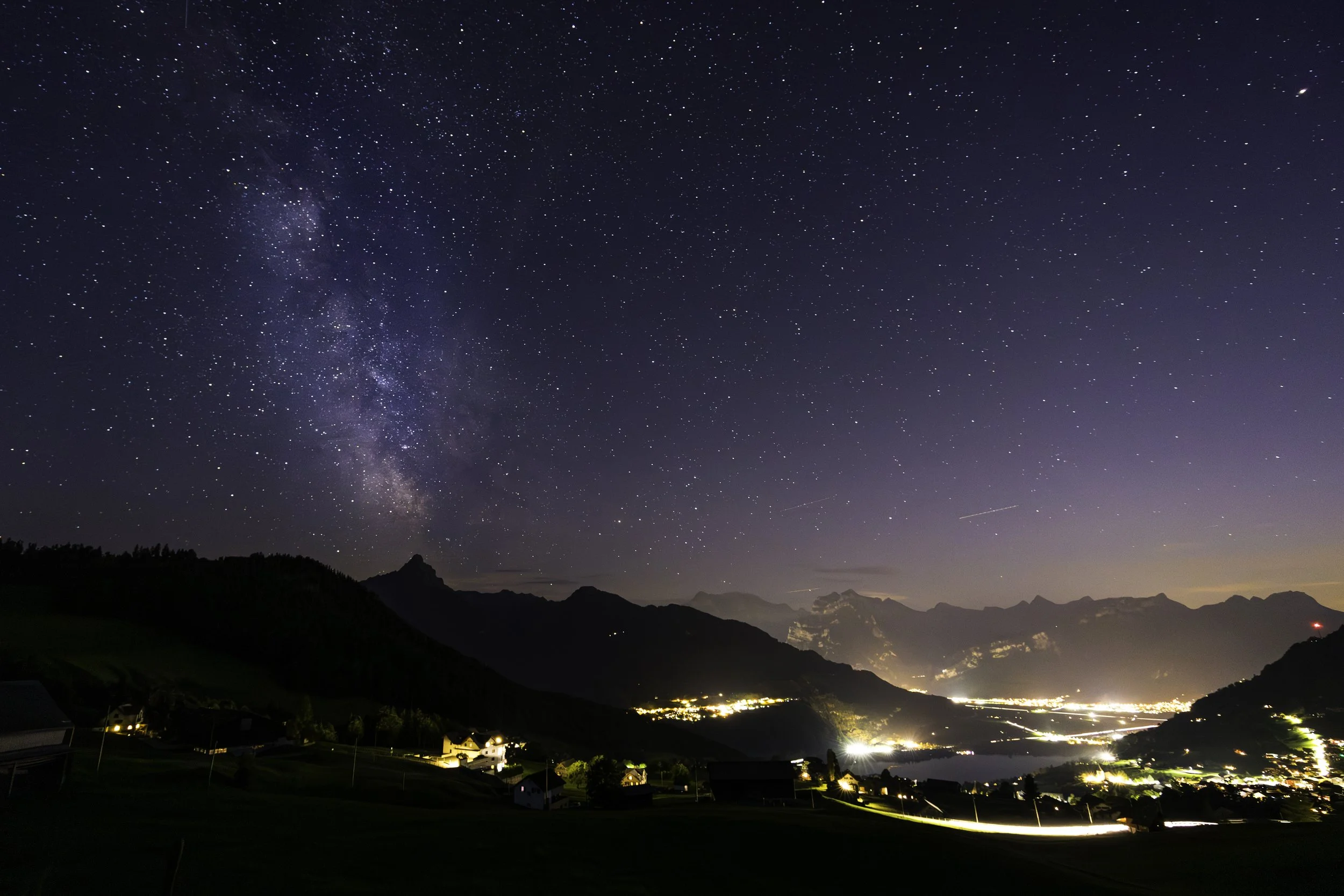Sternenklare Nacht über Bergen und einer kleinen Stadt mit leuchtenden Häusern und Straßen, im Himmel die Milchstraße sichtbar. Fotografie.