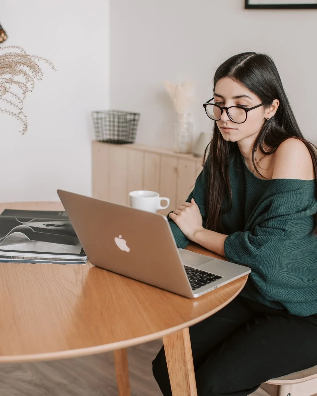 A young woman with long dark hair, glasses, and a green sweater sitting at a wooden table with a silver laptop open in front of her. There are notebooks, a white mug, and a background of a light-colored room with decorative items.