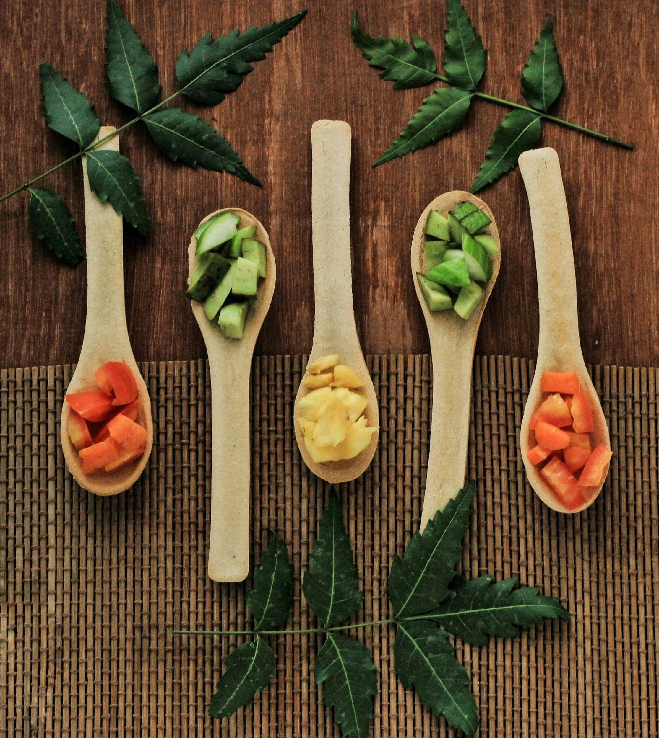Five wooden spoons filled with different chopped vegetables, arranged in a line on a bamboo mat with green leaves surrounding them, on a wooden table.