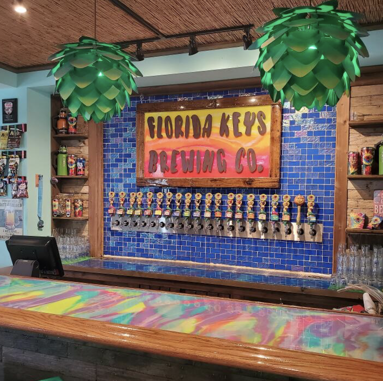 Bar area with a sign reading 'Florida Keys Brewing Co.', blue tiled wall, two large green leaf-shaped hanging lights, and tap handles adorned with colorful hats.