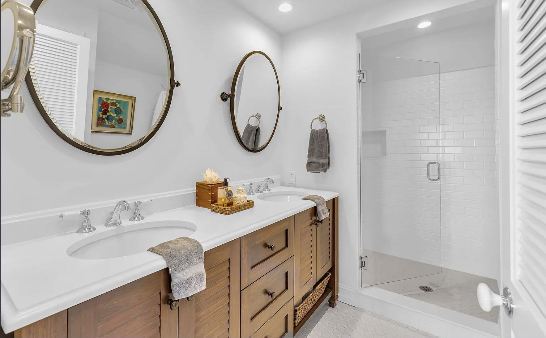 A bathroom with a double vanity with a white countertop and wooden cabinets, two oval mirrors with brown frames, and a glass-enclosed shower with white tiled walls.