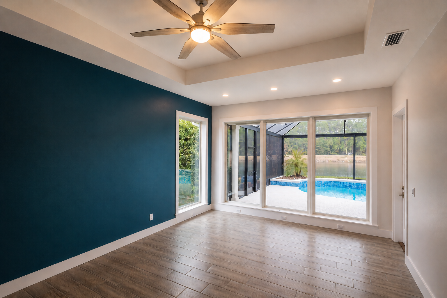 Empty living room with a blue accent wall, wood flooring, ceiling fan, and large windows showing a screened-in pool area and outdoor scenery.