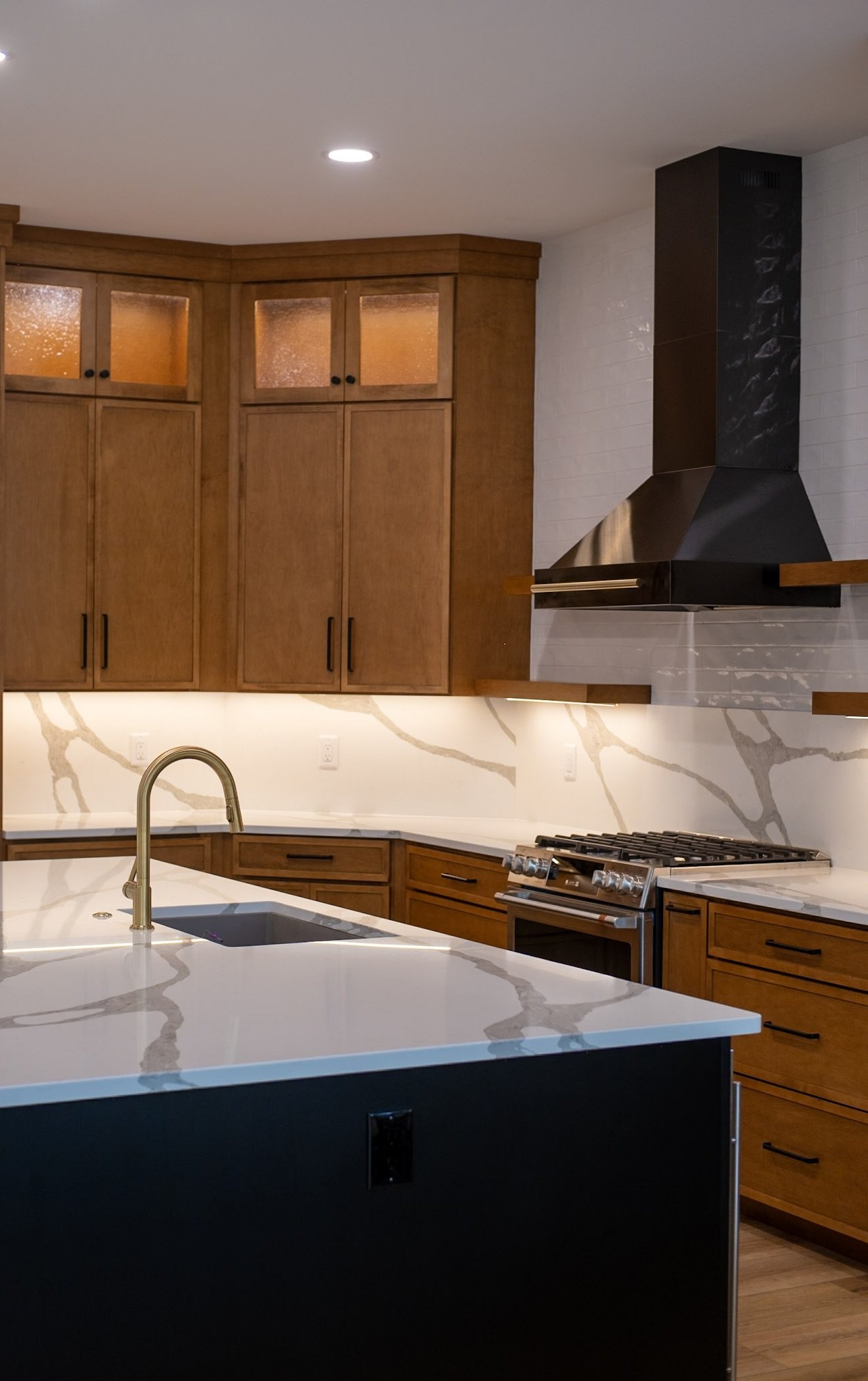 A modern kitchen with wooden cabinets, a white marble countertop with gray veining, stainless steel appliances, and a black range hood. The kitchen also features a kitchen island with a sink and a gold-colored faucet.