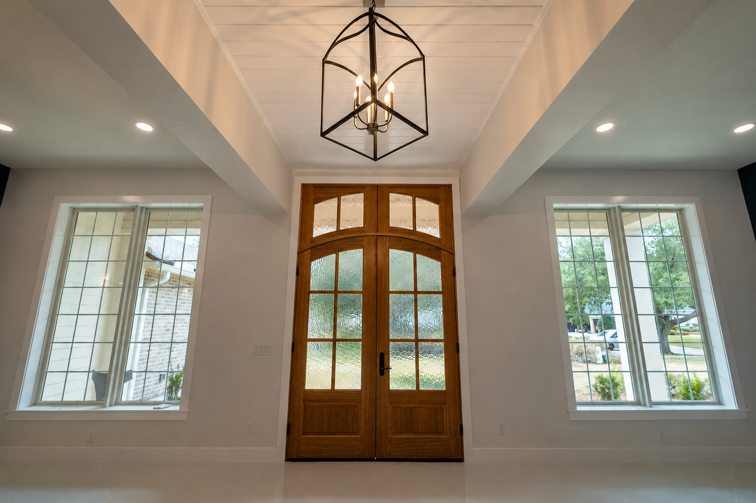 Interior view of a house entryway featuring a wooden double door with frosted glass panels, two large side windows with grid panes, a modern chandelier hanging from a white ceiling, and natural light coming through the windows.