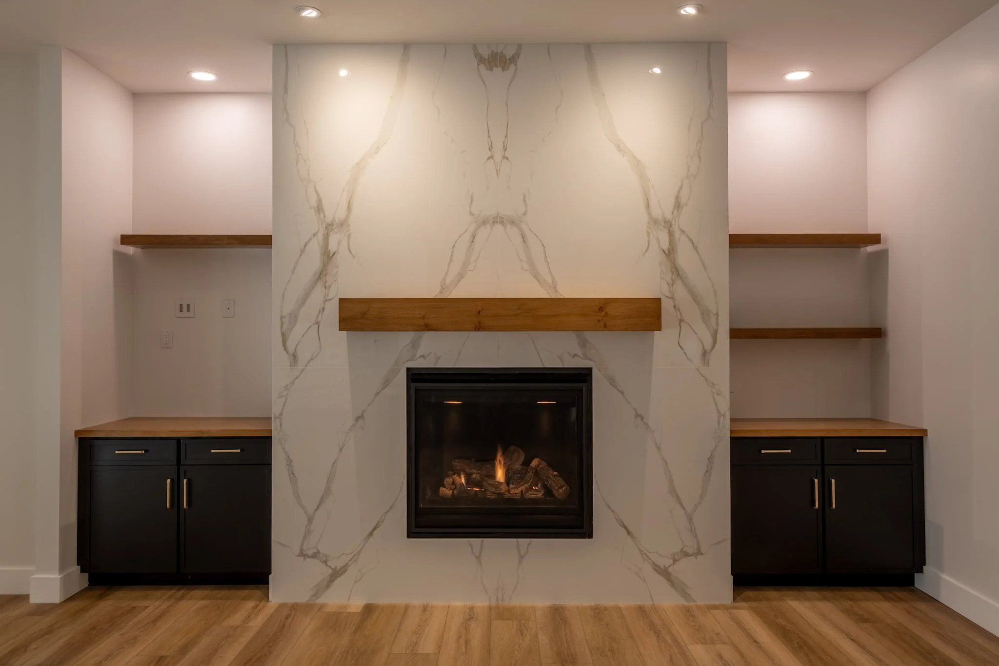A modern living room fireplace with marble surround, wooden mantel, and built-in shelves on each side, wooden flooring, recessed lighting, and a small fire burning inside the fireplace.