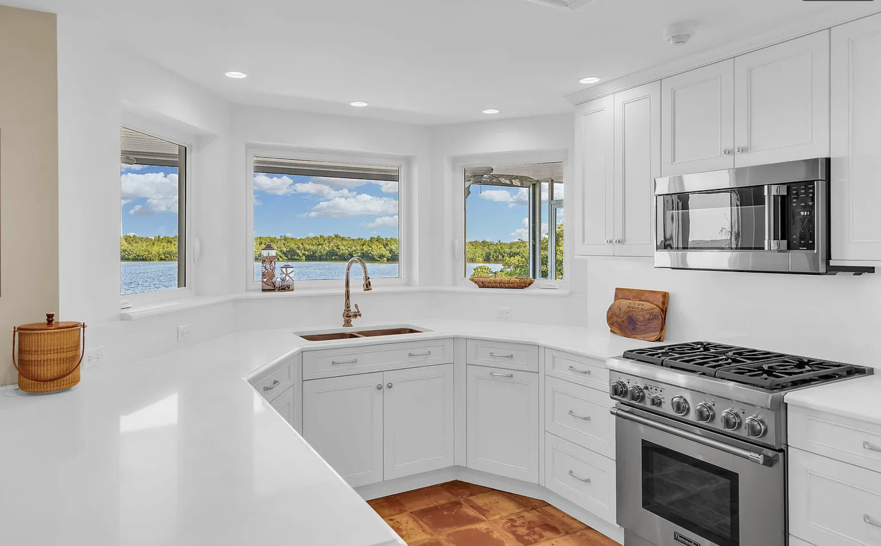 Bright kitchen with white cabinets and a large window overlooking a lake with trees and blue skies, a stainless steel stove and microwave, a wooden cutting board, and a copper faucet over a double sink.