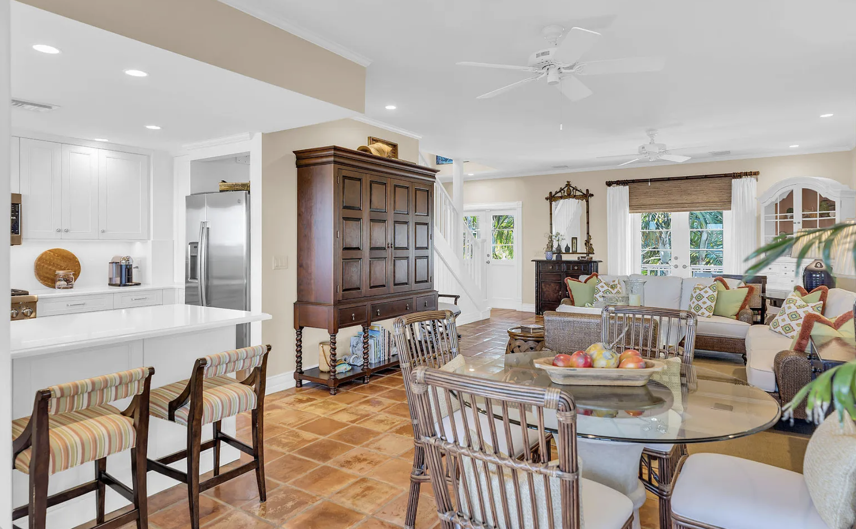 Living room with multiple seating areas, including a white sofa with colorful pillows, a wicker chair, and a glass dining table with chairs. There is a wooden cabinet and a mirror, large windows with white curtains, and a brick tile floor.