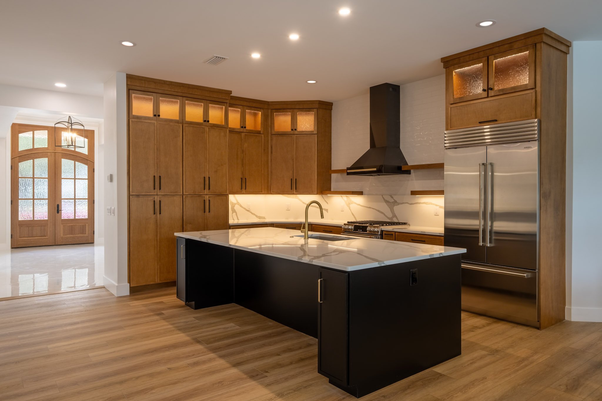 Modern kitchen with wooden cabinets, stainless steel refrigerator, black island with white marble countertop, and a white brick backsplash.