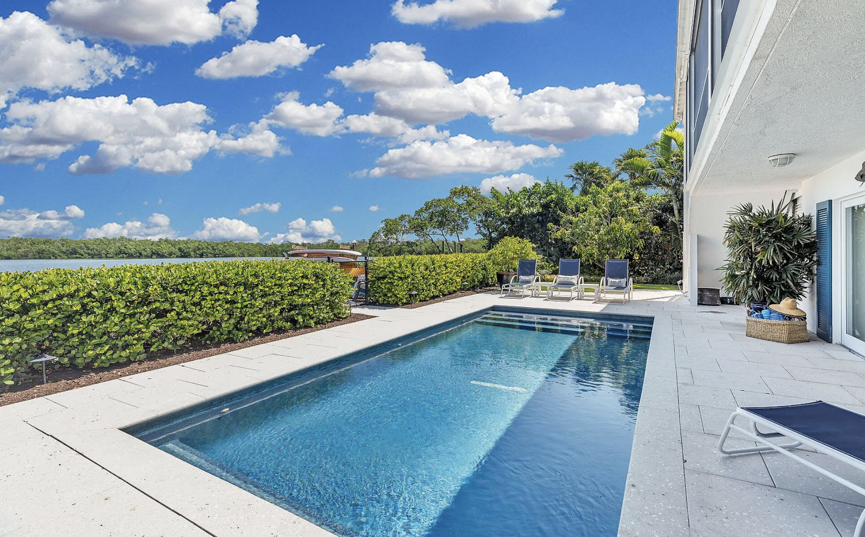 A backyard swimming pool with clear blue water, surrounded by a white stone deck, overlooking a green hedge and a body of water under a blue sky with scattered clouds.