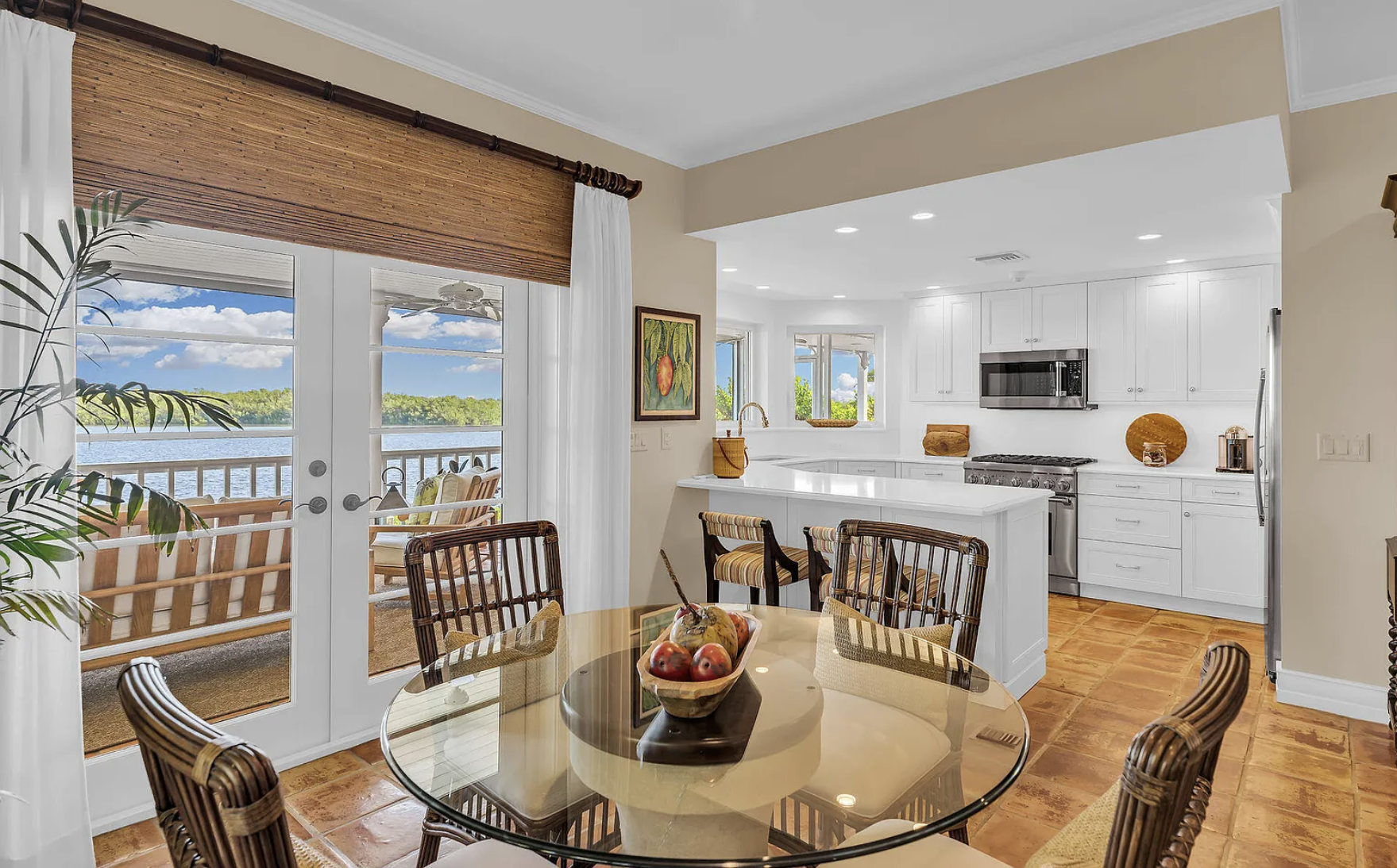 A bright open kitchen and dining area with a view of a lake outside. The kitchen features white cabinetry, stainless steel appliances, and an island. The dining table has a bowl of apples, and there are chairs with wicker seating. French doors with g