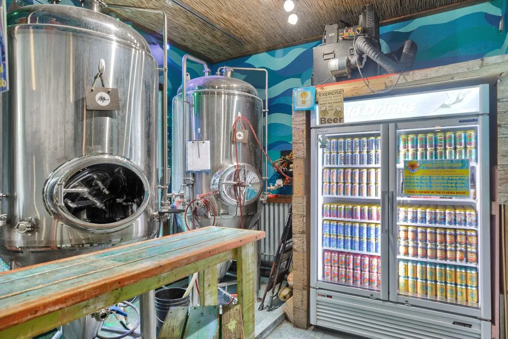 Inside a brewery or beverage production area with large stainless steel fermenters or brew tanks, a wooden workbench, and a refrigerated display case filled with cans of beer.