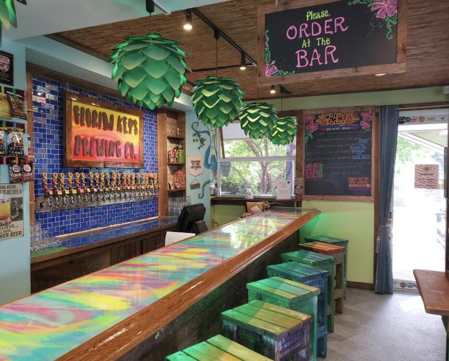 Interior of a colorful bar with a rainbow-colored counter, green leaf-shaped hanging lamps, and a blackboard menu on the wall. There is a sign that says "Please Order at the Bar" and a row of beer taps on a blue tiled wall.