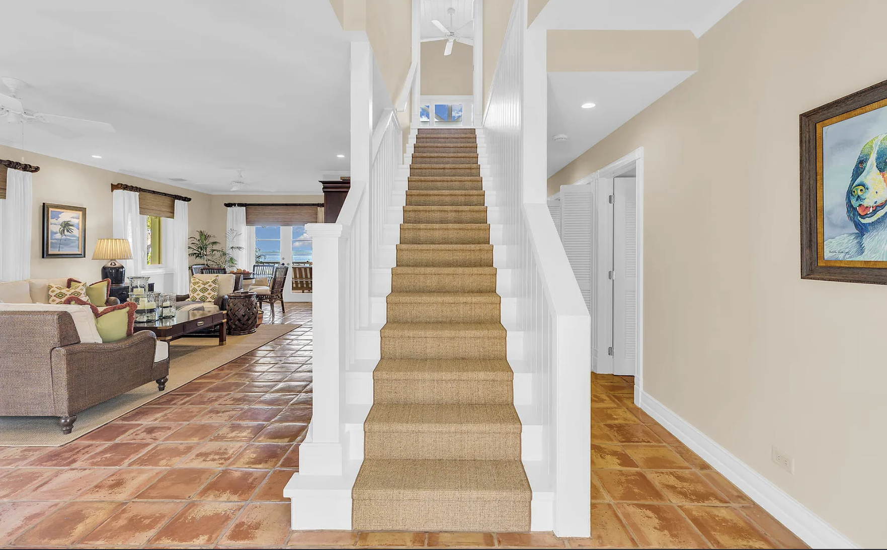 Interior view of a home with a staircase in the center, beige walls, tiled flooring, part of a living room with sofa, chairs, and artwork on the walls.