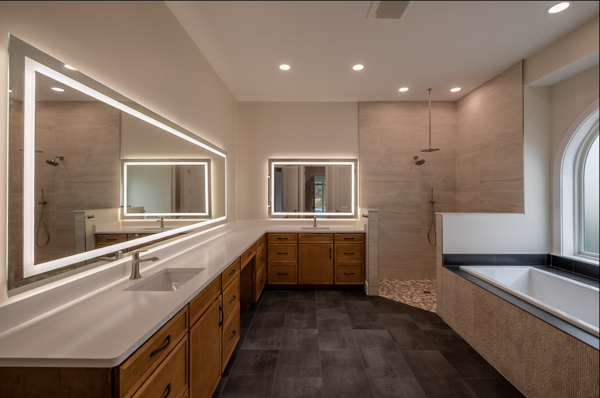 Modern bathroom with a double vanity with wooden cabinets, large illuminated mirrors, a bathtub, and a walk-in shower with beige tile walls and a window.