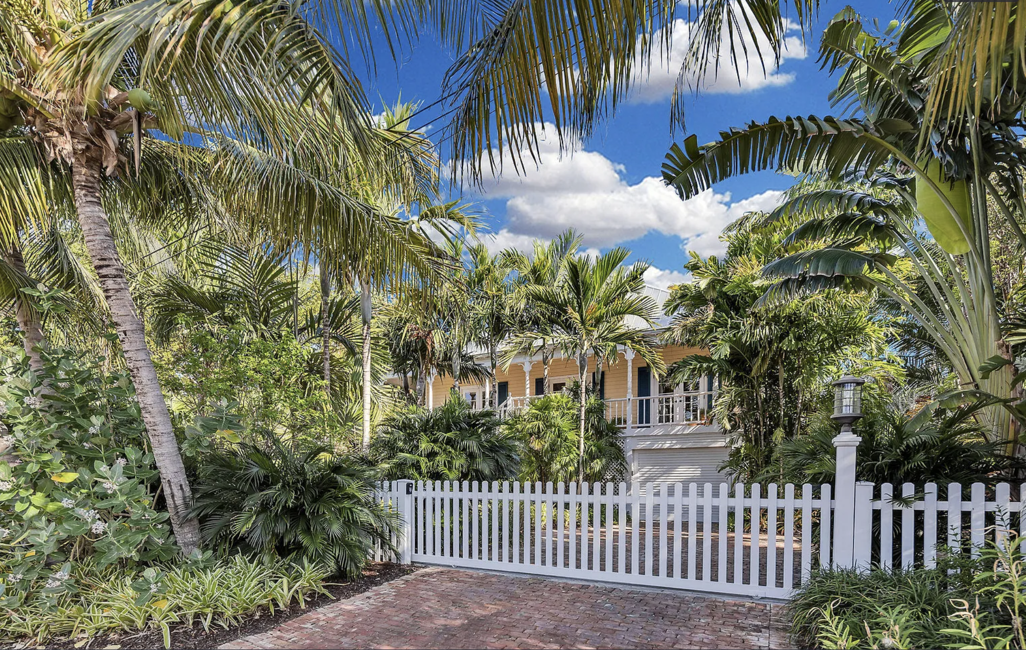 Tropical garden with lush green palm trees, a white picket fence, brick pathway, and a two-story house with a porch and white railing, under a partly cloudy blue sky.