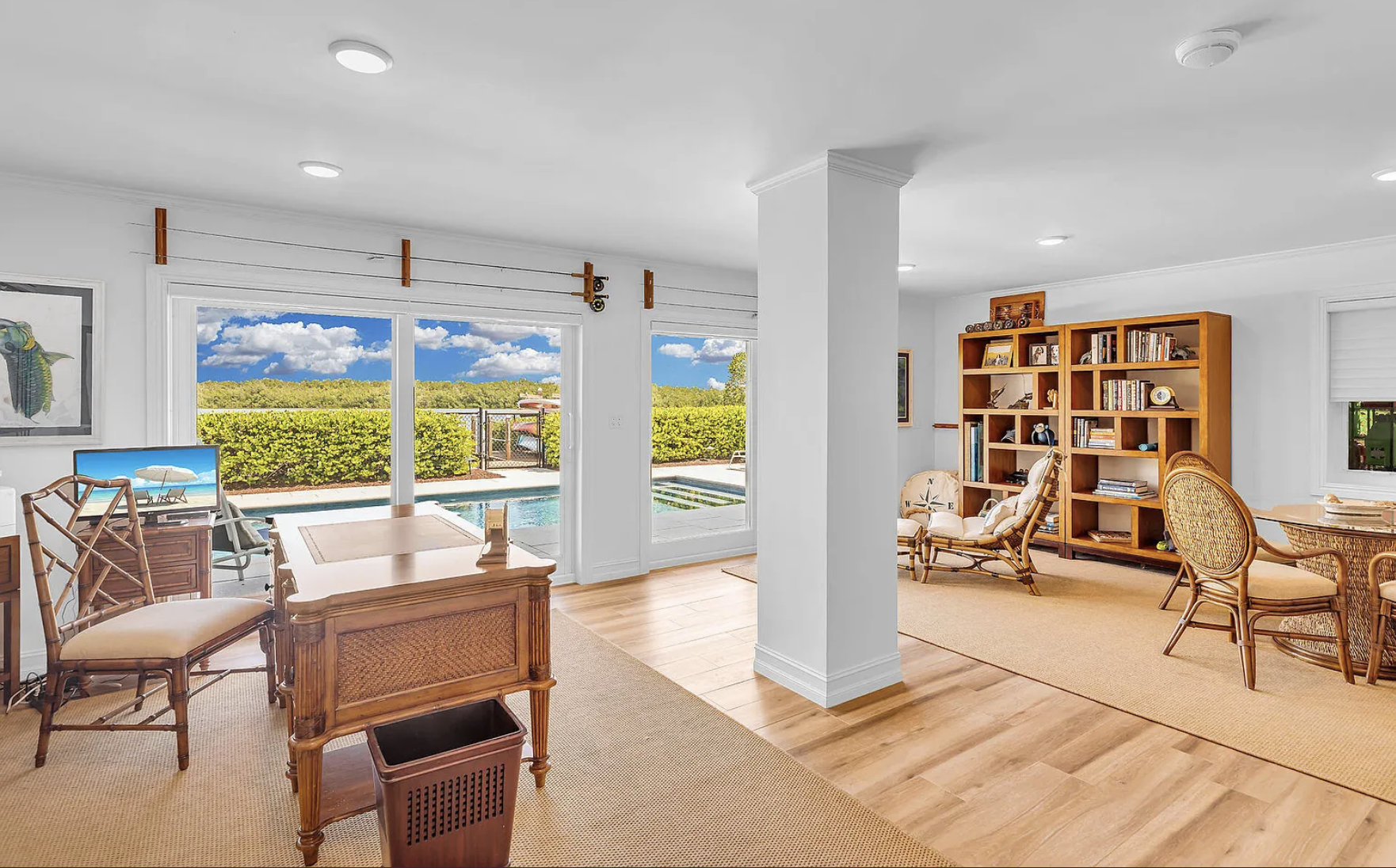 Living room with a view of a pool and backyard through large sliding glass doors, featuring wooden and wicker furniture and a wooden bookshelf.