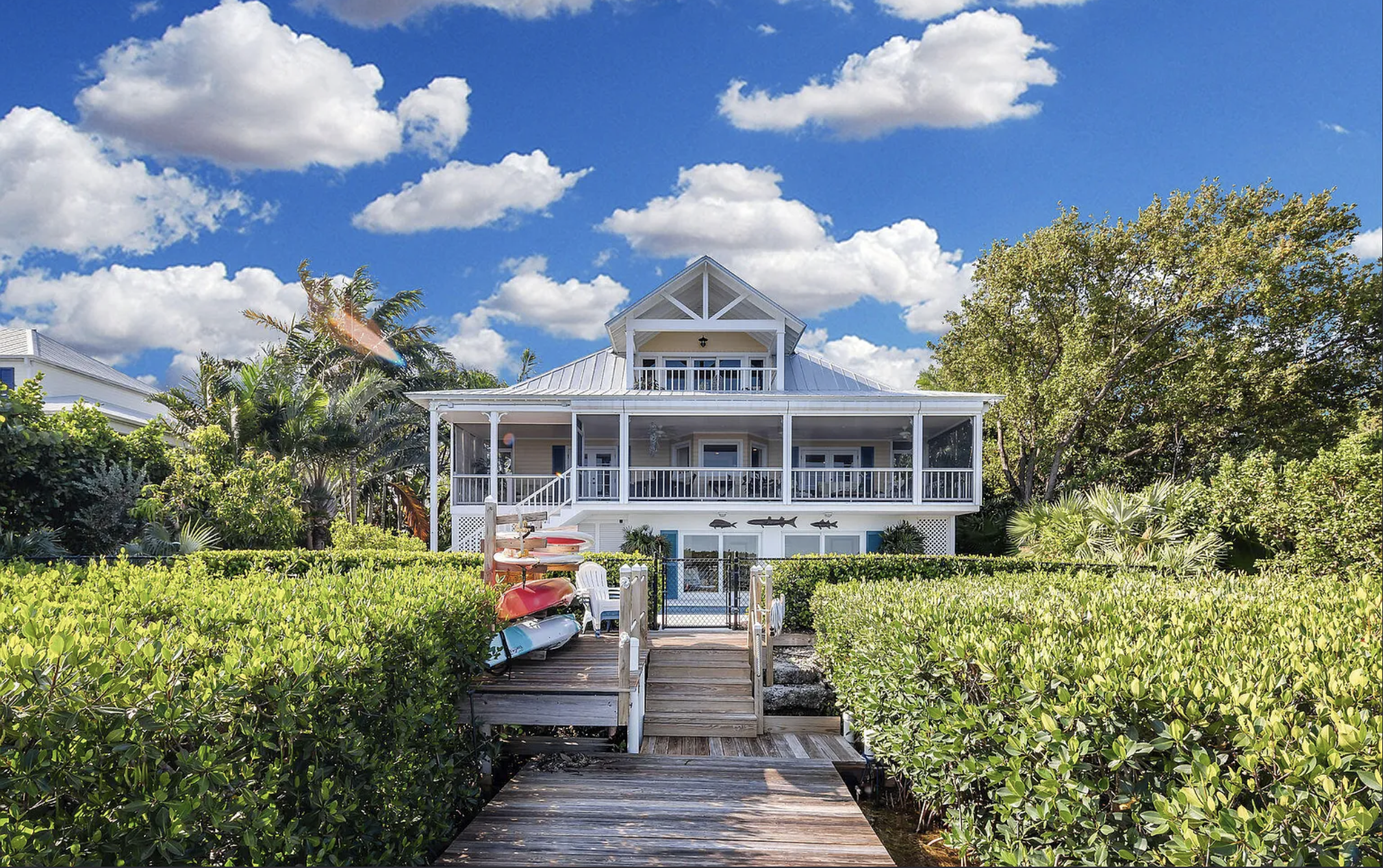 A white seaside house with multiple balconies, surrounded by lush green plants and trees, under a blue sky with scattered white clouds.