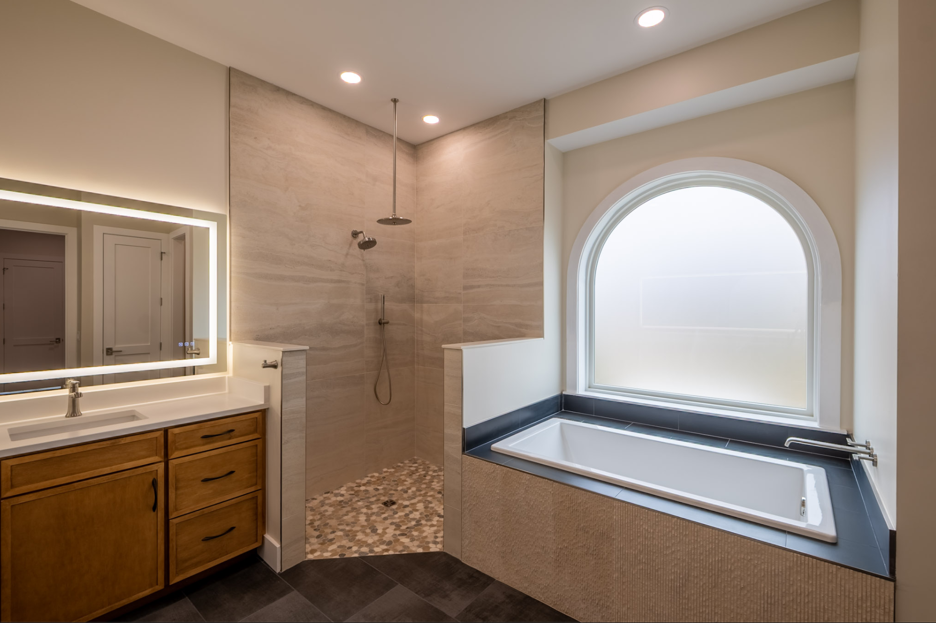 Modern bathroom featuring a large bathtub beneath a frosted window, a walk-in shower with beige tiles, a wooden vanity with a backlit mirror, and recessed ceiling lights.