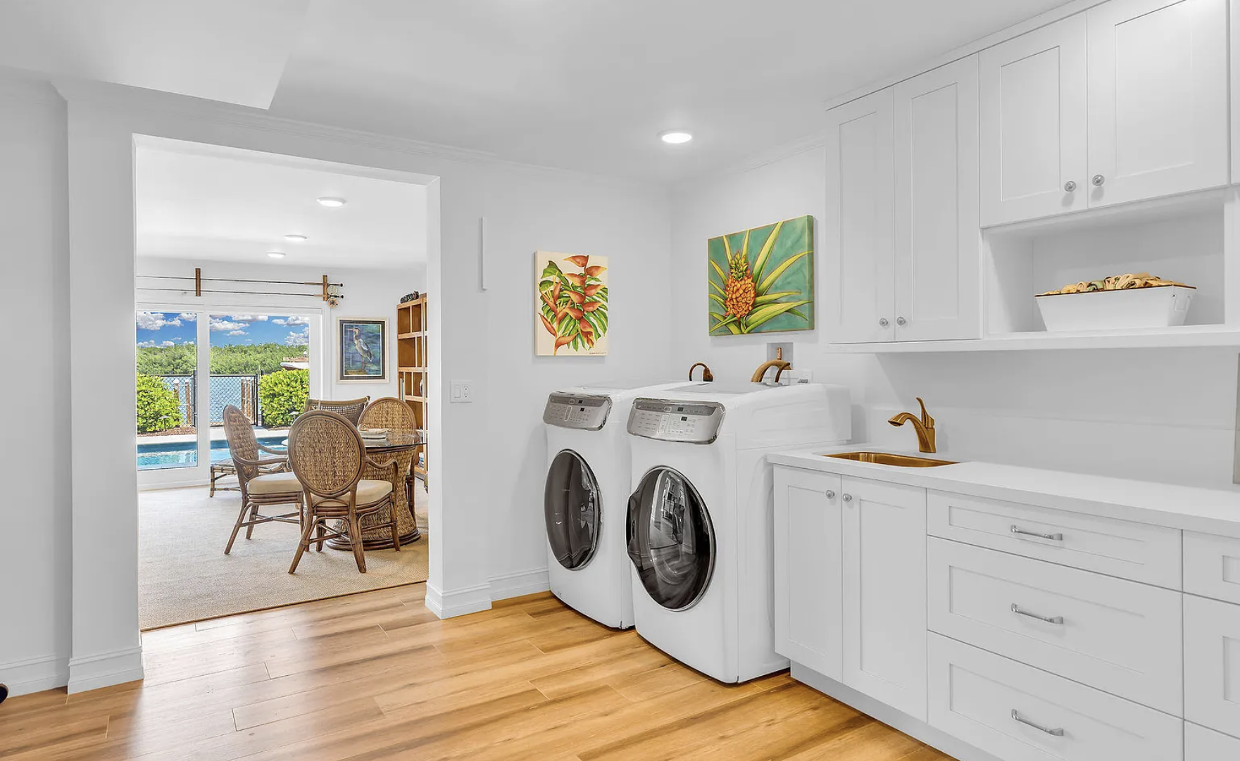 Laundry room with washing machine and dryer, white cabinets, gold faucet, small sink, and colorful pineapple-themed paintings on the wall. Visible through doorway is a dining area with a table, wicker chairs, sliding glass door, and pool outside.