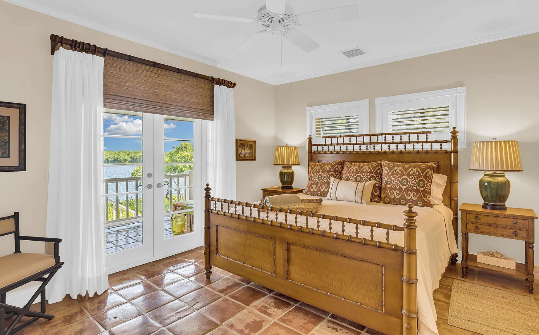 A cozy bedroom with a wooden bed, nightstands, and lamps, featuring large windows and a door leading to a balcony with a scenic water view.