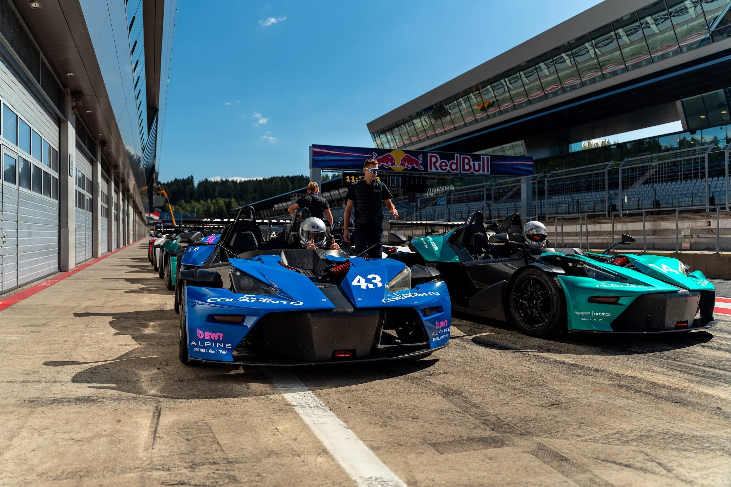 Zwei KTM X-Bow Rennwagen mit helmtragenden Fahrern startbereit in der Boxengasse des Red Bull Ring, im Hintergrund das Boxengebäude mit Red Bull Schriftzug