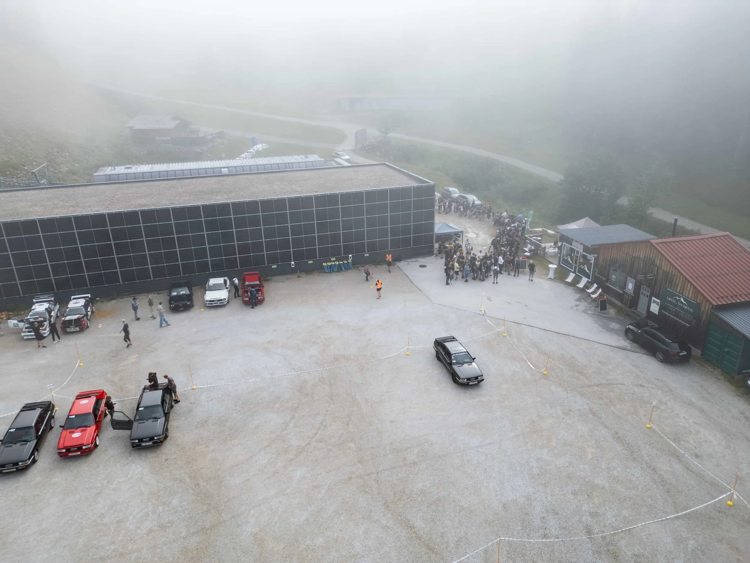 Luftaufnahme eines Auto-Events bei Nebel vor einer Gondel und Berghütten in den österreichischen Bergen.