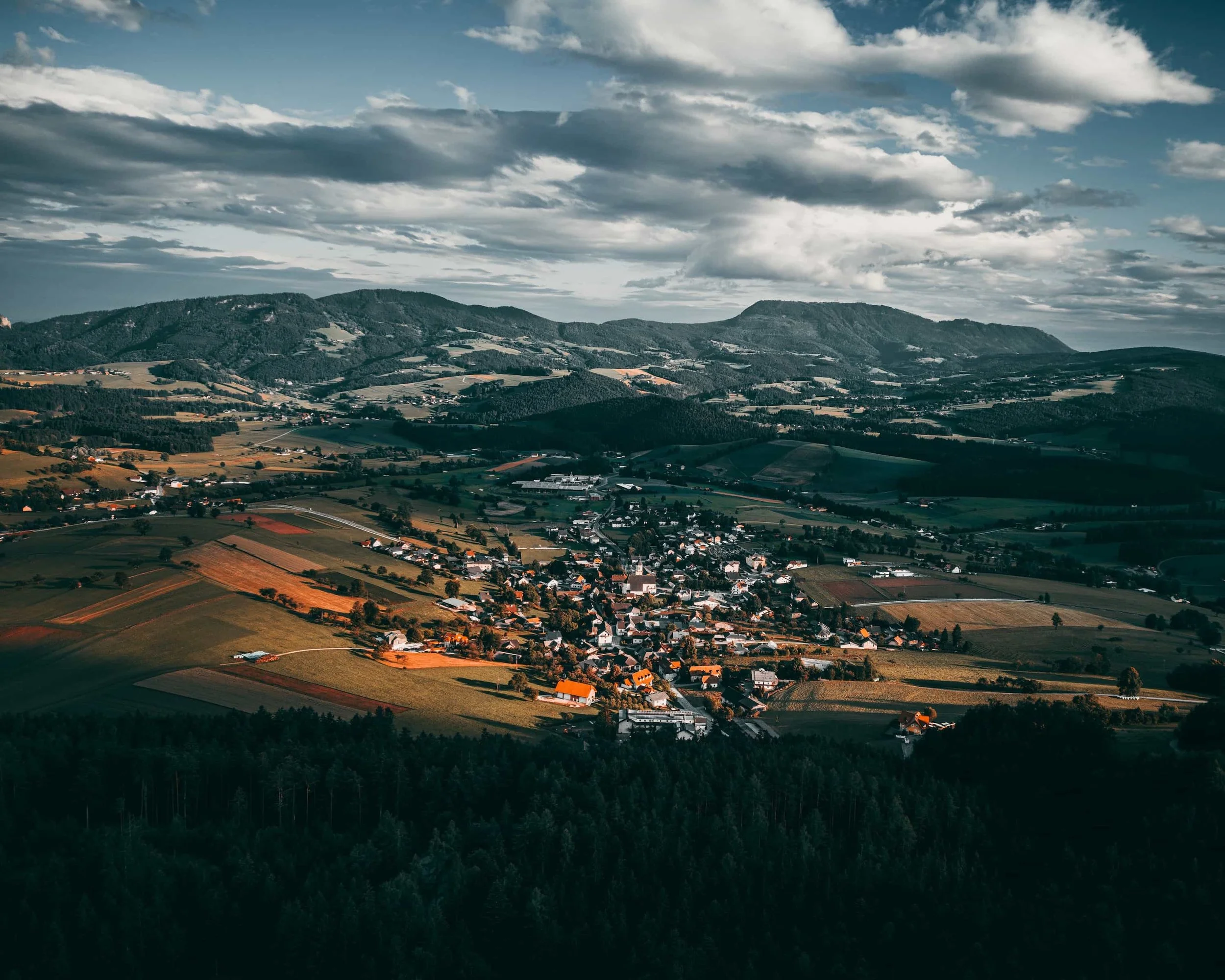 Drohnenaufnahme einer Ortschaft im österreichischen Alpenvorland mit Bergen und Feldern im warmen Abendlicht.