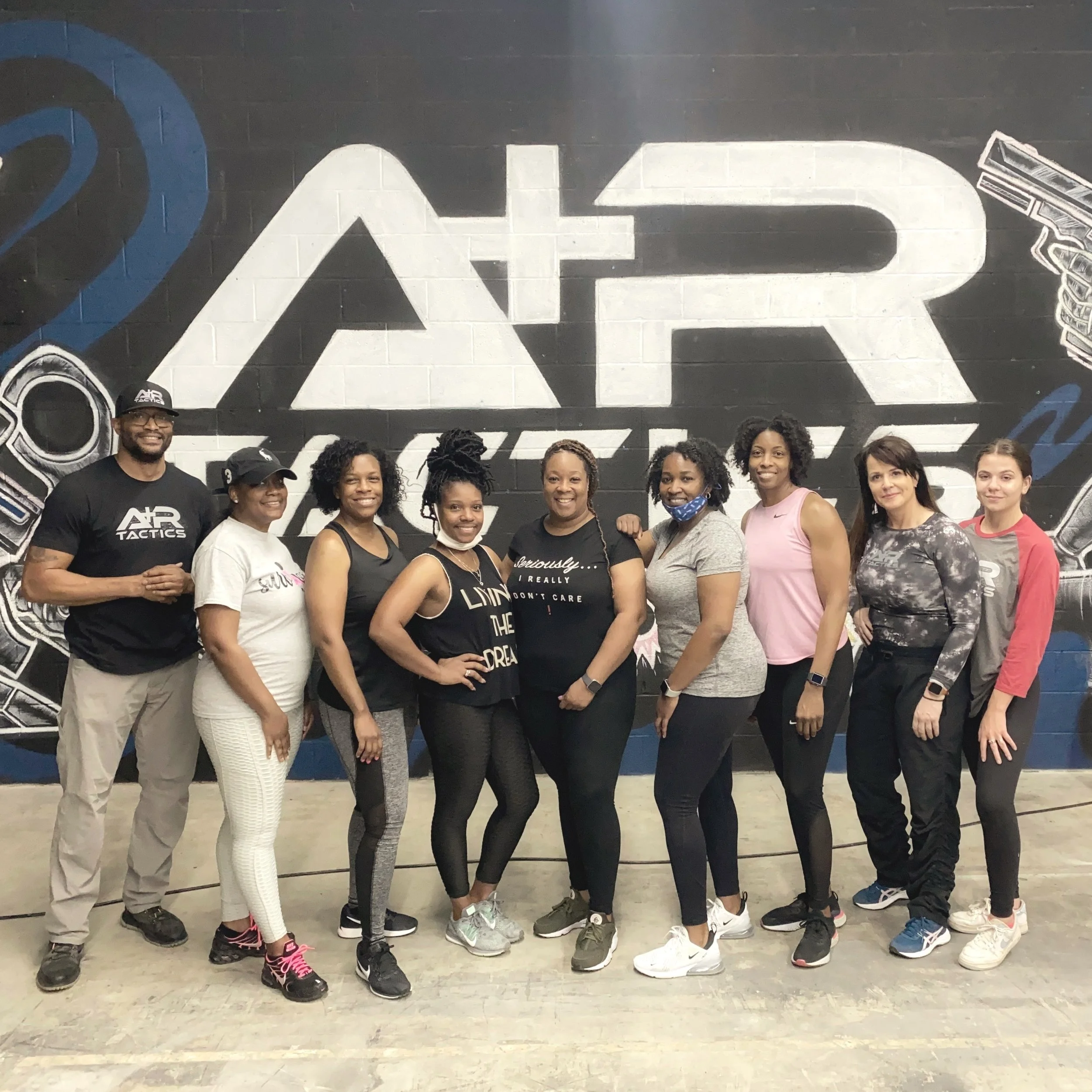 Group of people posing in front of A&R Tactics logo on a wall. Corporate events, self-protection and team-building at A+R Tactics shoot house firearms training.