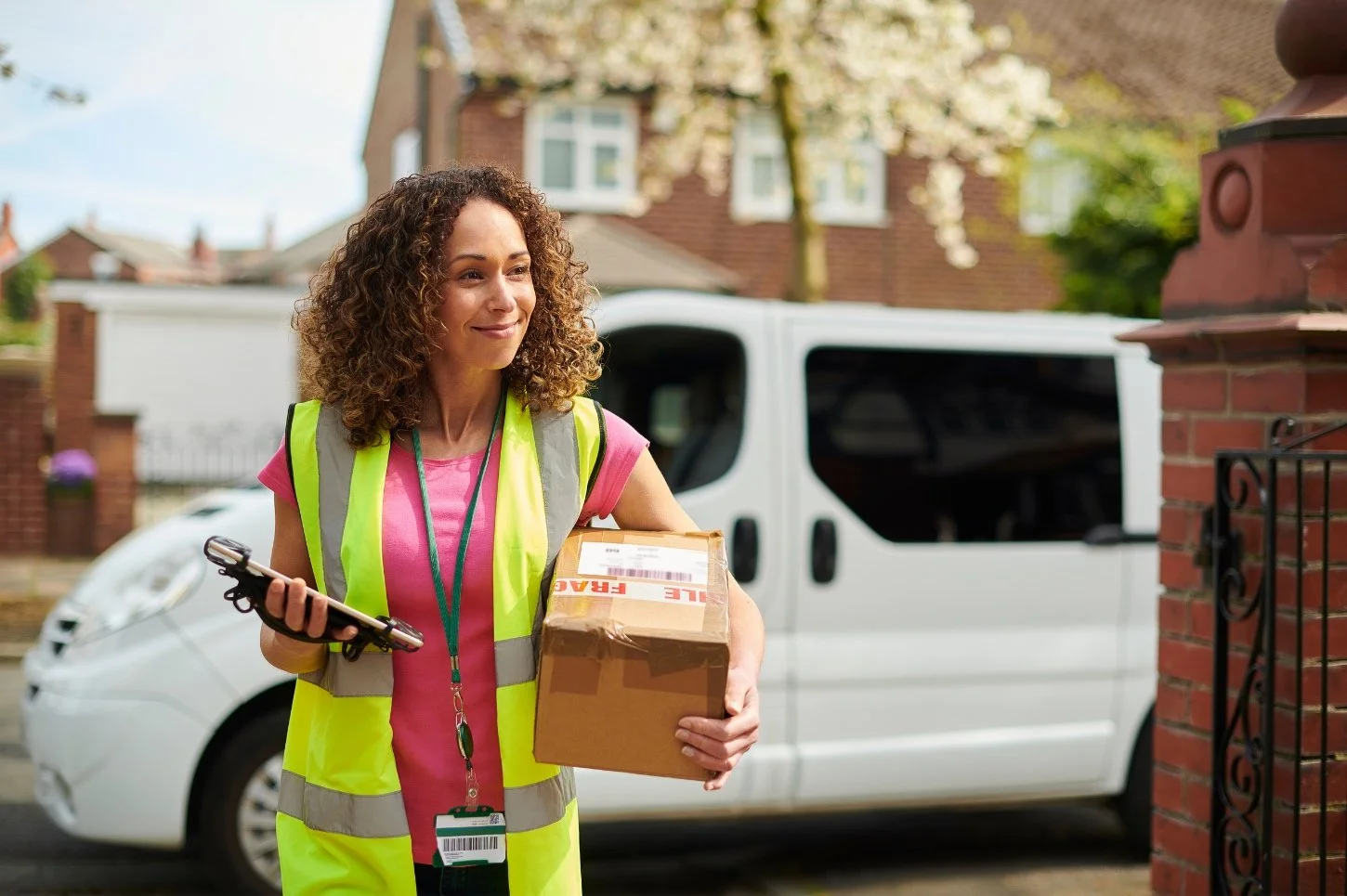 Delivery person wearing a high-visibility vest holding a package and electronic device, standing in front of a white delivery van in a residential area.