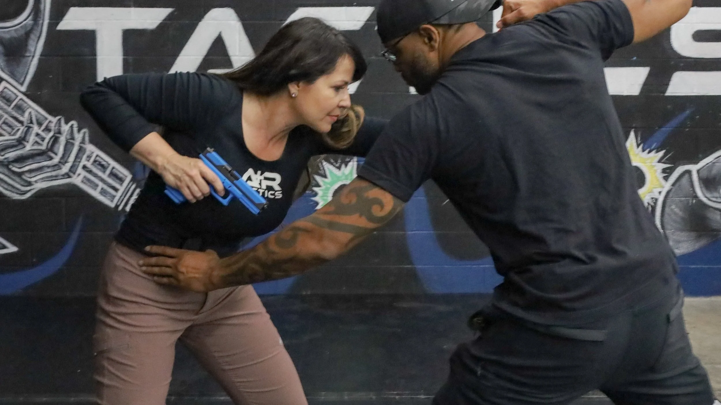 Two people practicing self-defense techniques with a blue training gun, indoors. Corporate events, self-protection and team-building at A+R Tactics shoot house firearms training.