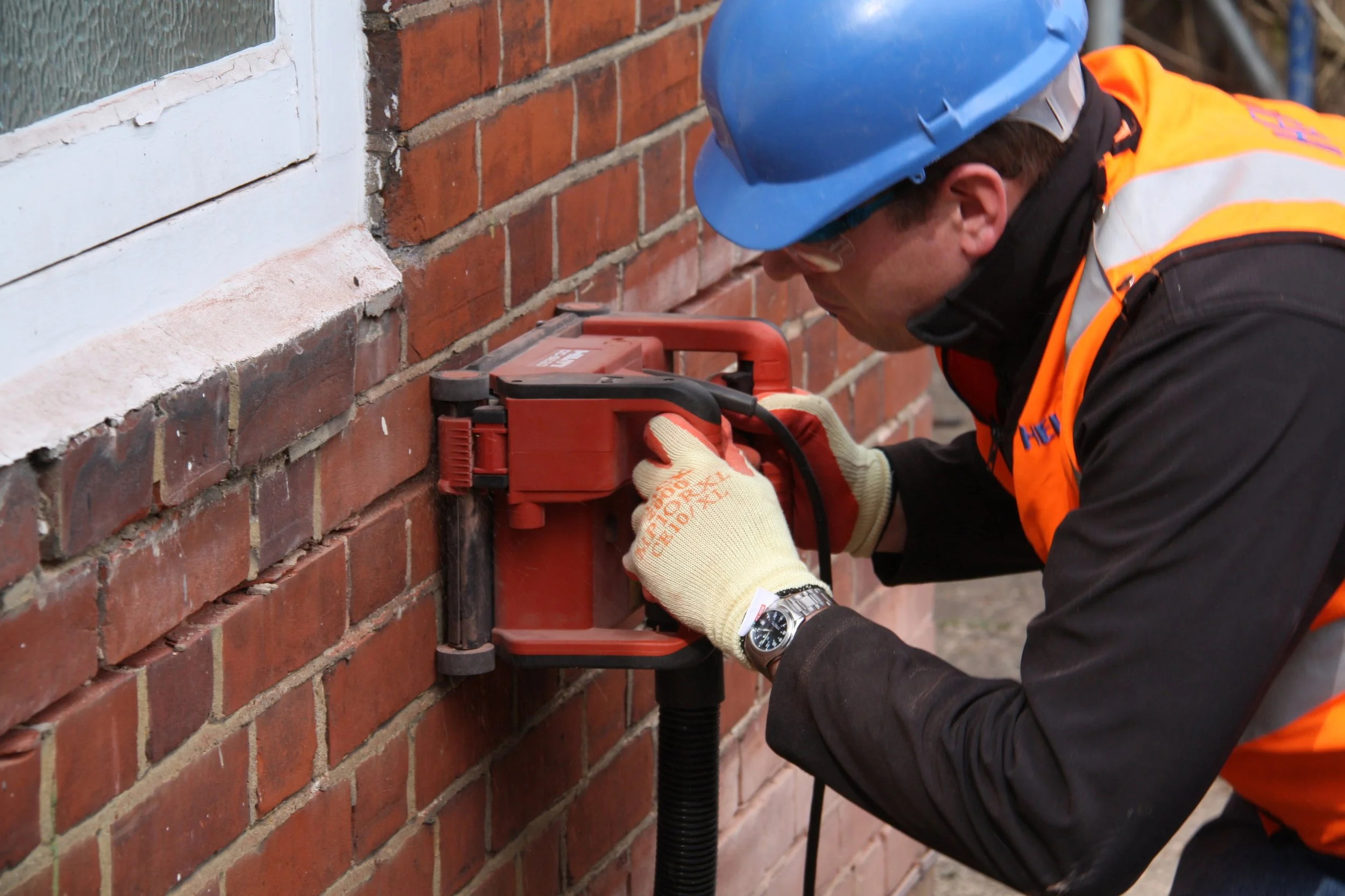 Construction worker using a red power tool for drilling into a brick wall, wearing a blue hard hat, orange safety vest, and gloves.