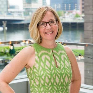 A woman with shoulder-length blond hair and glasses smiling outdoors with a city waterfront in the background, wearing a green patterned sleeveless dress.