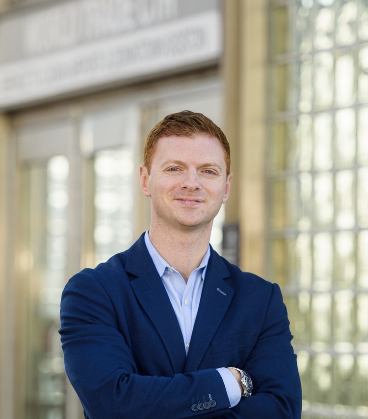 A young man with red hair and a light skin tone wearing a navy blazer and light blue shirt, smiling with arms crossed, standing inside a building with large windows.