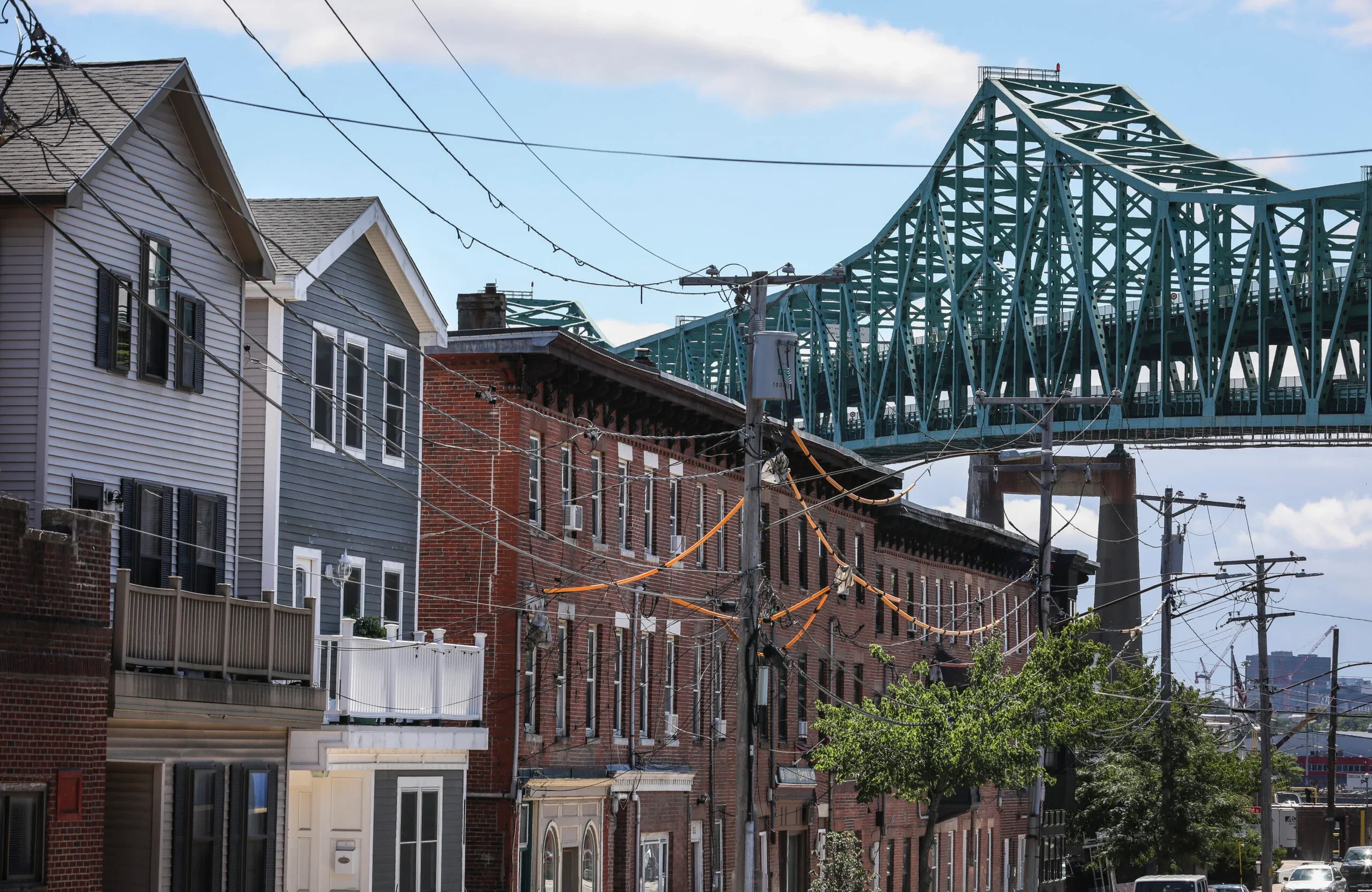 Cityscape showing a row of residential buildings with colorful siding and brick facades, power lines, street trees, and a large teal steel bridge in the background.