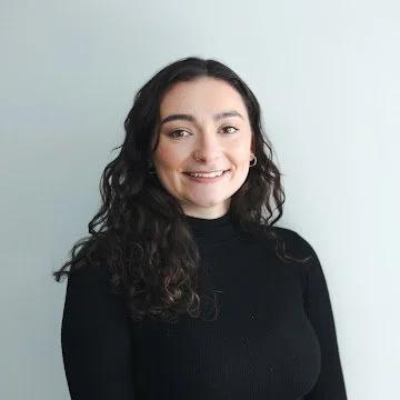 A young woman with dark wavy hair wearing a black long-sleeve top, smiling against a light-colored background.