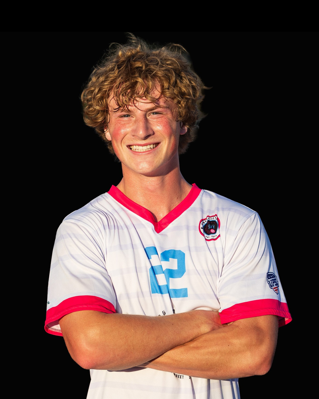 A young man with blond curly hair standing with arms behind his back, wearing a black soccer jersey with light blue accents, the number 4 in pink, and a logo on the chest