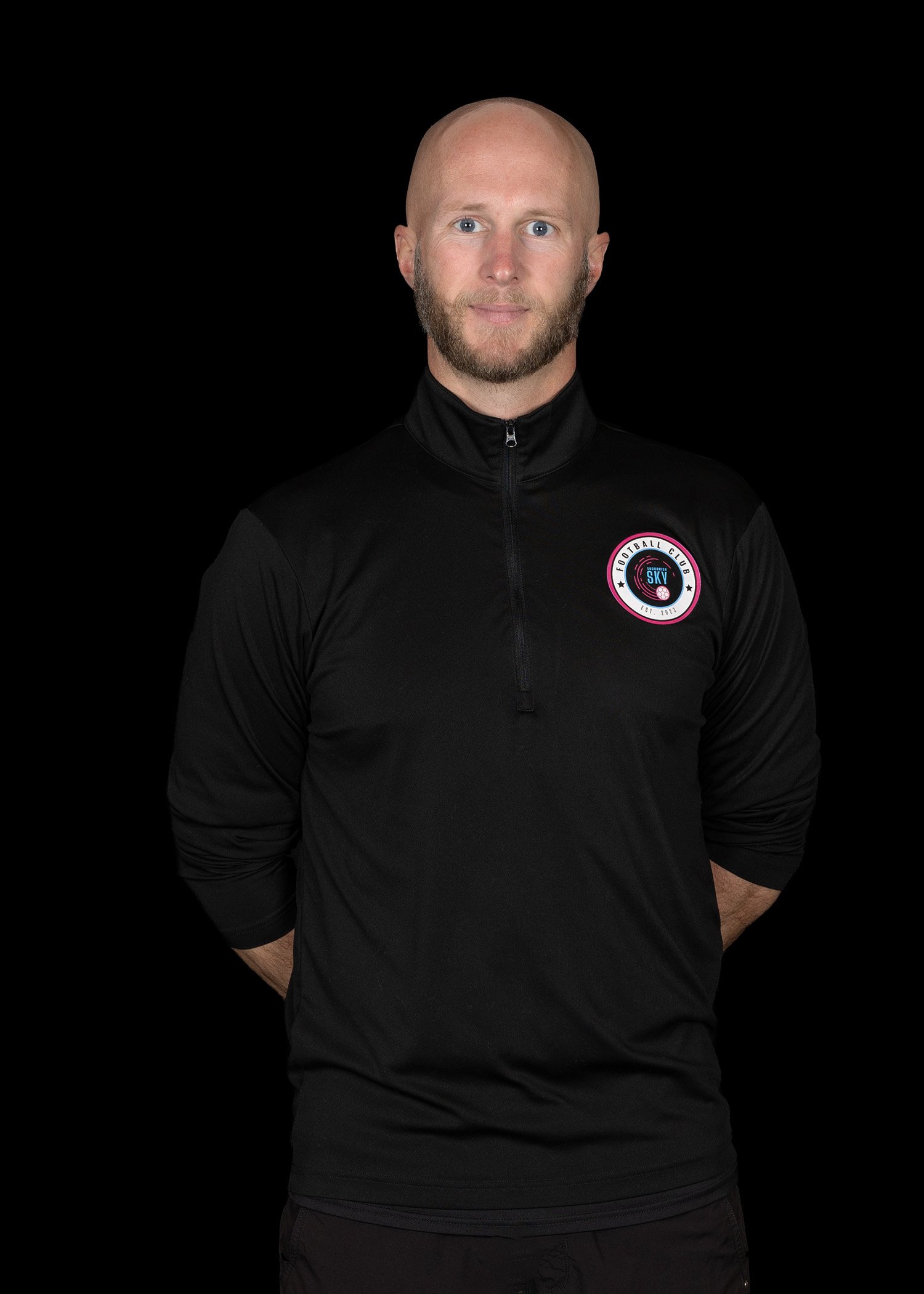 A smiling man with a bald head and beard, wearing a black polo shirt with logos, standing against a dark and colorful abstract background.