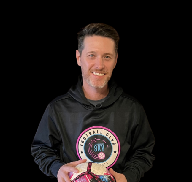 Smiling man with short dark hair and a beard, wearing a black hoodie with a pink and white logo in front, standing against a textured dark wall.