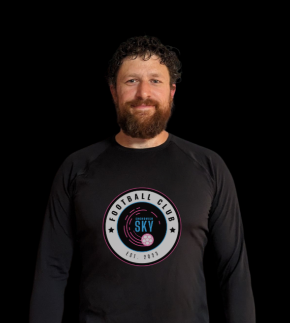 Man with curly hair and beard smiling, wearing a black shirt with "Snohomish Sky" logo and a pink soccer ball graphic.