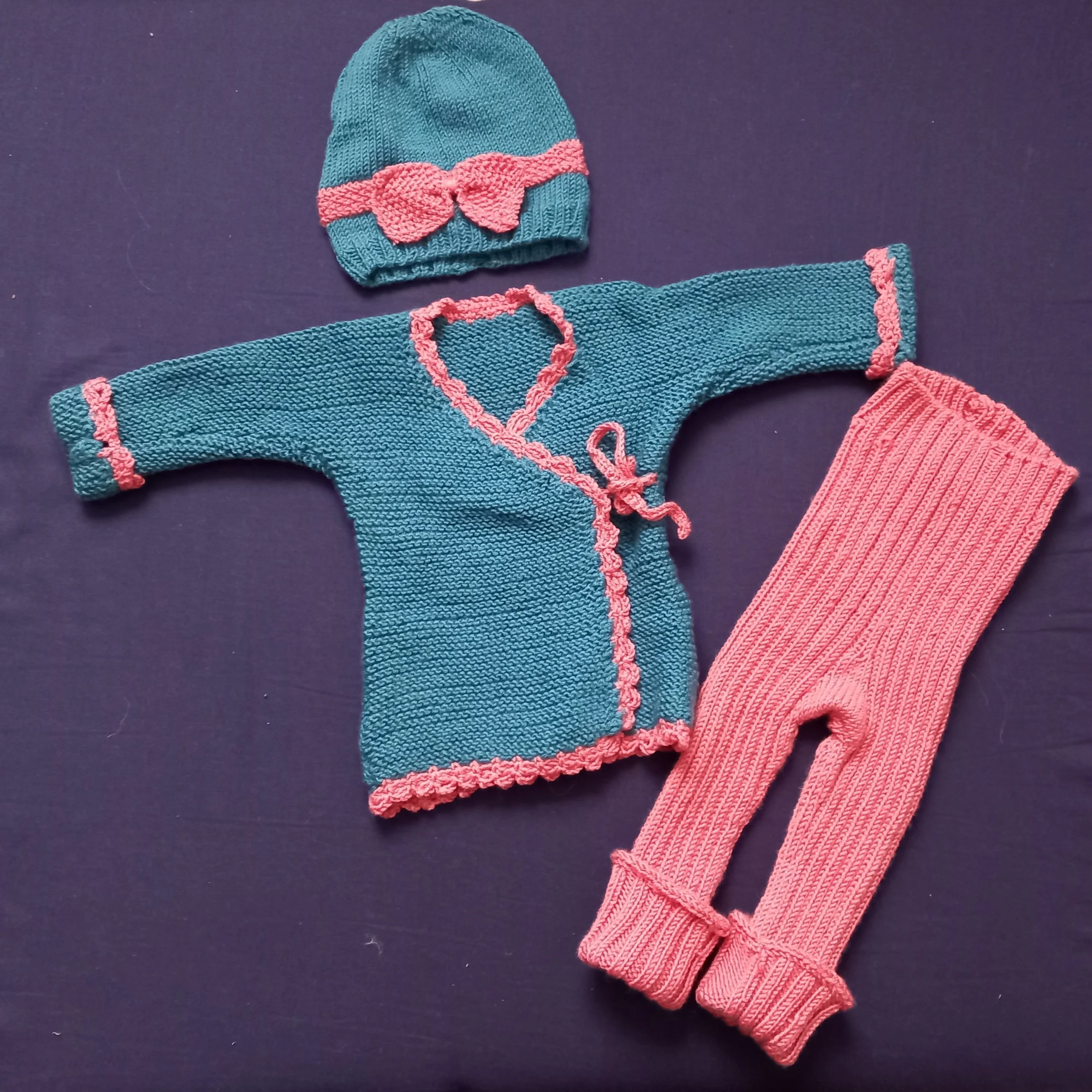 Kleine Kleidung für ein Baby, bestehend aus einer blauen Strickjacke mit pinken Details, einem pinken Hut mit pinker Schleife und pinken Sonnenbrillen, und einer Packung pinker Strumpfhosen.